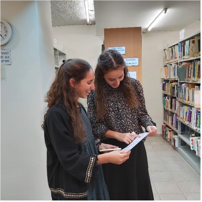 משחוק בספרייה- 2 סטודנטיות קוראות ספר על רקע מדפי הספרייה